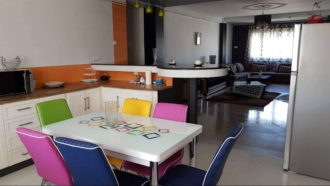 A modern kitchen and dining area features a white table surrounded by colorful chairs in green, blue, yellow, and pink. The kitchen includes stainless steel appliances and a bar counter. In the background, a living area showcases a sofa with patterned cushions and large windows allowing natural light.