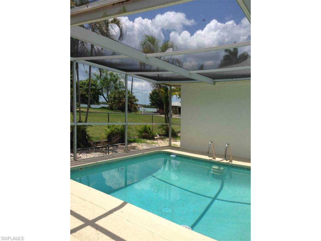 A pool area is enclosed by a screen cage, featuring clear water that reflects the sky. Green grass and trees are visible in the background, alongside a well-maintained lawn. Sunlight brightens the space, creating a relaxed atmosphere for enjoyment.