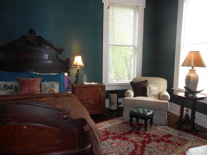 The room features dark teal walls that provide a rich backdrop for elegant antique furnishings. A queen-sized bed is accented with decorative pillows. A cozy club chair and a small footstool invite relaxation, while light filters through large windows adorned with blinds.