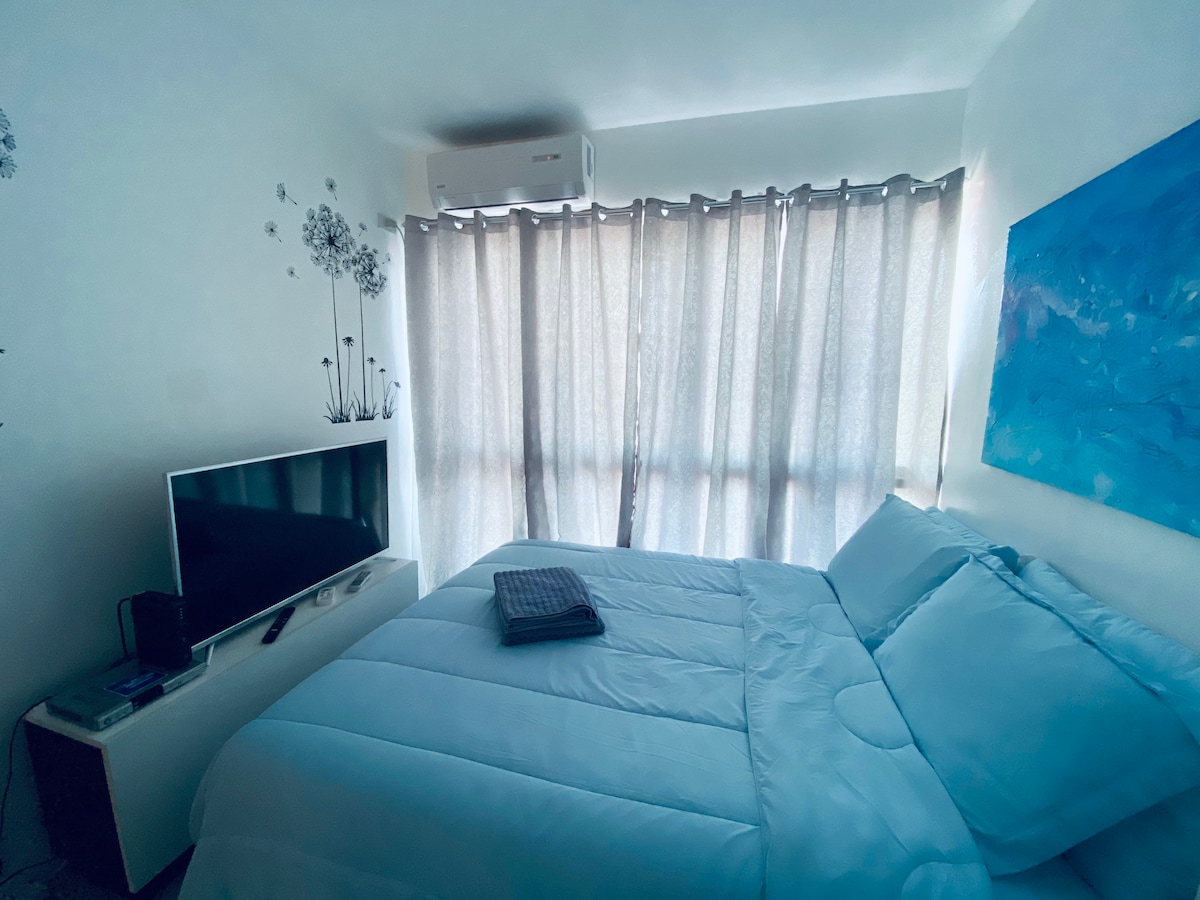 A cozy bedroom features a spacious bed with crisp white linens, complemented by decorative pillows. A television is positioned on a minimalist dresser. Light filters through sheer curtains, creating a soft glow in the room, while floral wall decals add a touch of elegance.