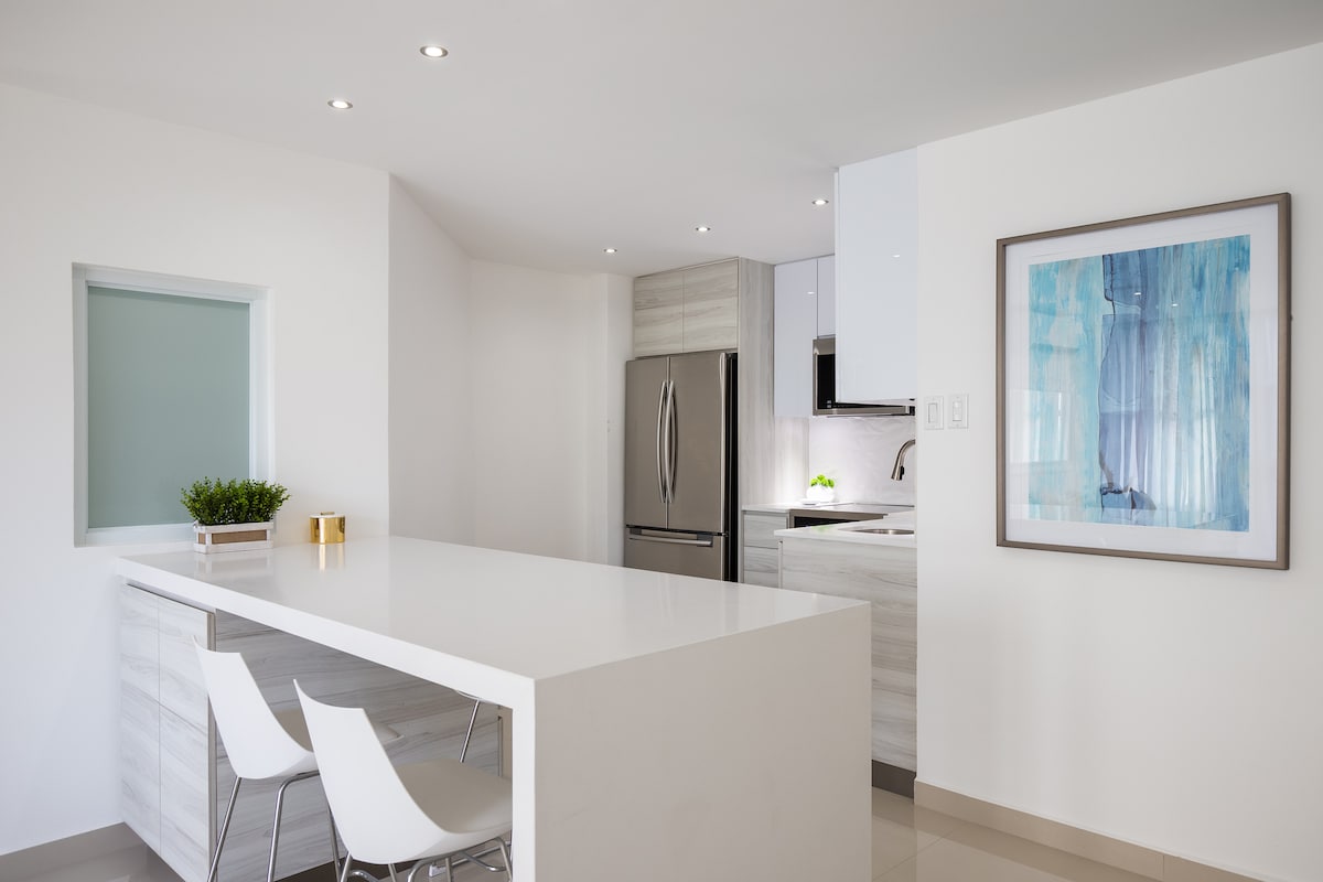 A modern kitchen features a sleek white countertop with two minimalist chairs. Stainless steel appliances, including a refrigerator, are positioned against light cabinetry. A colorful artwork adds a touch of elegance to the bright, open space, enhanced by recessed lighting above.