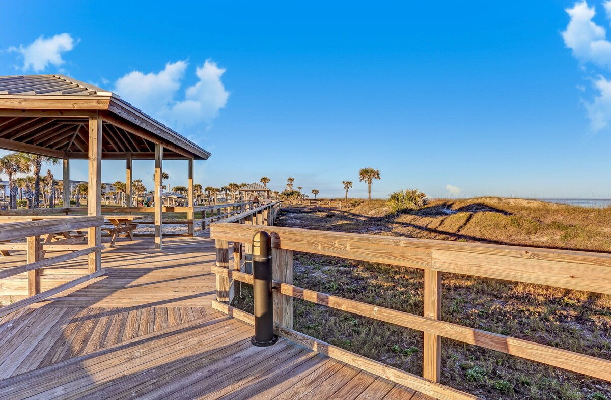 A wooden deck extends toward the ocean, featuring a partially covered seating area. Natural grass and palm trees are visible in the background, under a clear blue sky with few clouds. A trash receptacle stands at the edge of the deck.