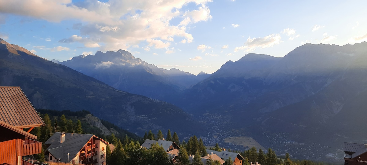 A panoramic view captures mountains and valleys under a clear sky, with gentle sunlight illuminating the peaks. The landscape features a mix of green forests and rustic chalets dotting the foreground, creating a serene connection with nature.