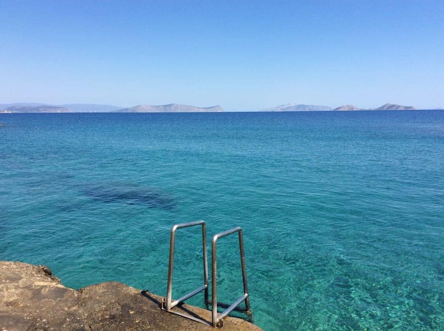A clear view of the sea is showcased, featuring calm turquoise waters that extend to distant islands. A metal ladder is positioned on a rocky edge, providing access to the pristine water, under a bright blue sky.