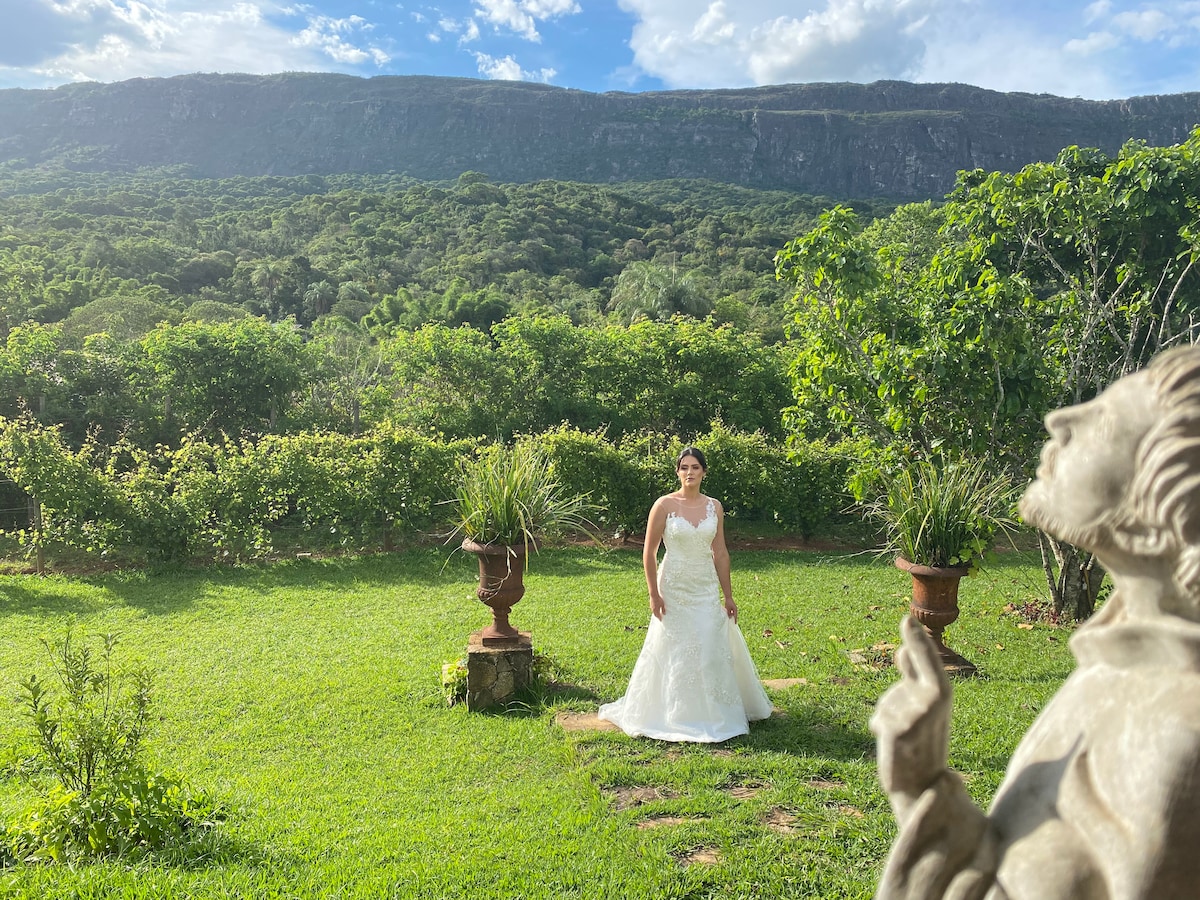 An outdoor garden space features lush greenery and potted plants. The background showcases a scenic view of the Serra de São José mountains under a partly cloudy sky. A figure in a white dress stands gracefully amidst the vibrant landscape, enhancing the tranquil setting.