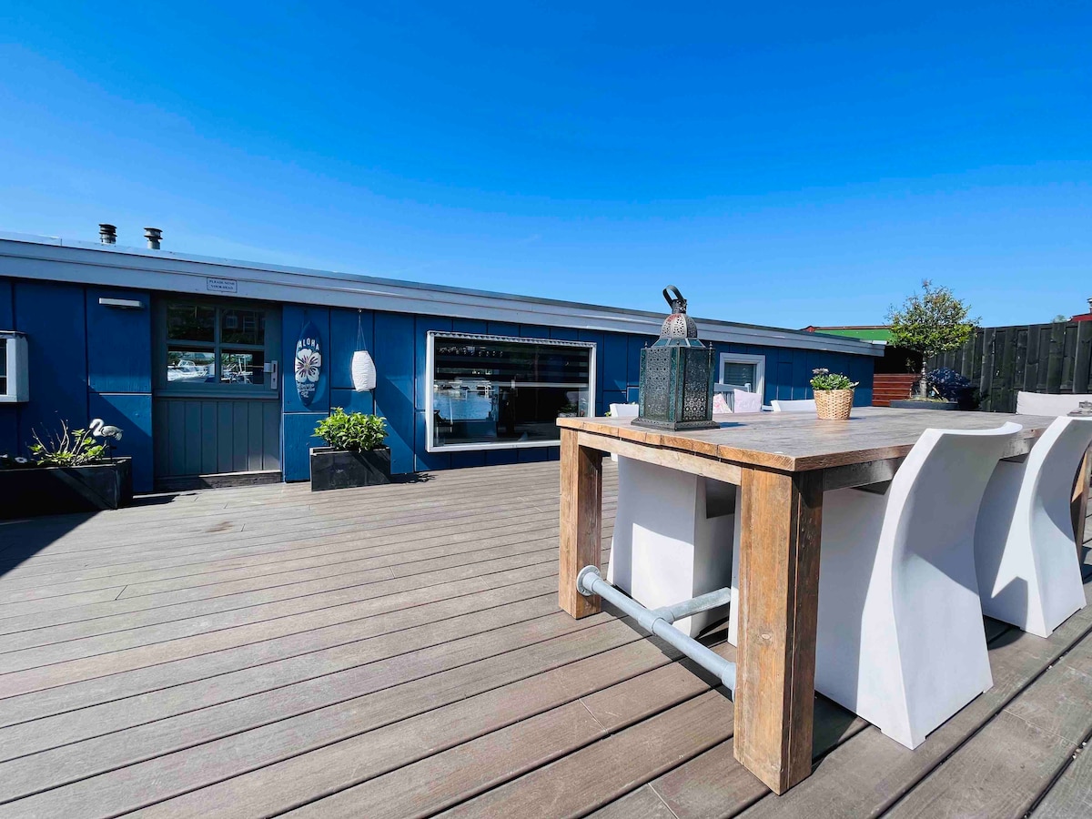 A spacious outdoor terrace features a wooden dining table accompanied by white chairs. Planters with greenery add to the inviting atmosphere. The blue exterior of the boat is complemented by clear skies, providing a bright and open space for guests to relax.