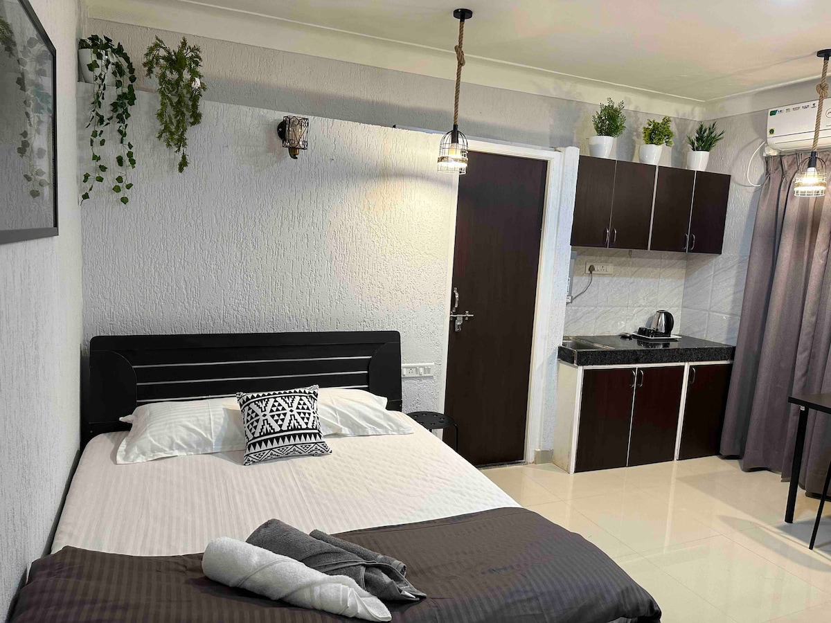 A cozy studio room features a comfortable queen-sized bed, adorned with decorative pillows and a soft blanket. A kitchenette with dark cabinetry and a kettle is visible in the background. Potted plants are placed above the cabinets, and light fixtures add a modern touch.