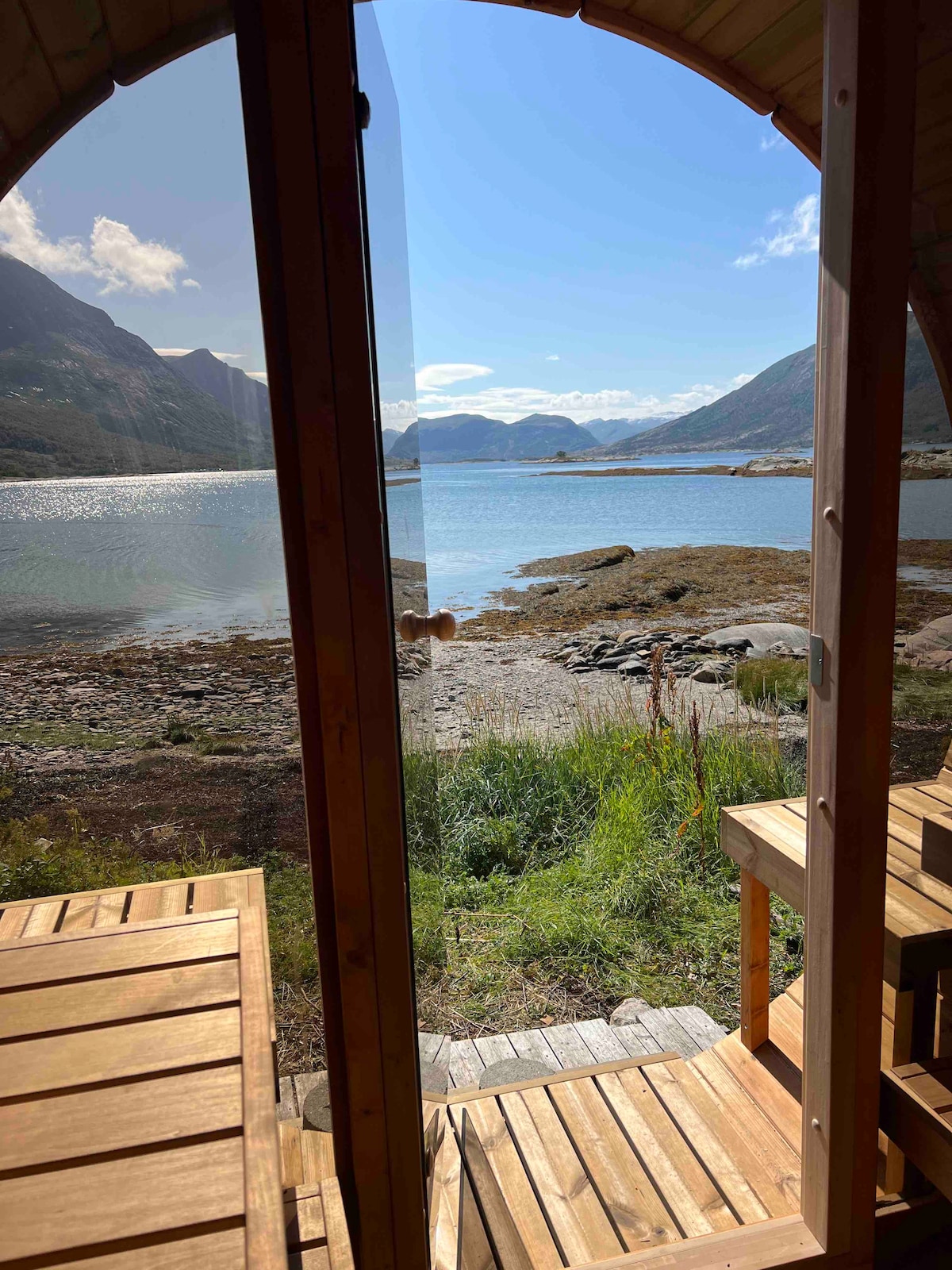 Glass doors provide a clear view of the tranquil fjord, where calm waters reflect the surrounding mountains. A wooden deck features seating, inviting relaxation while enjoying the natural scenery. The landscape includes rugged terrain and greenery, enhancing the serene atmosphere.