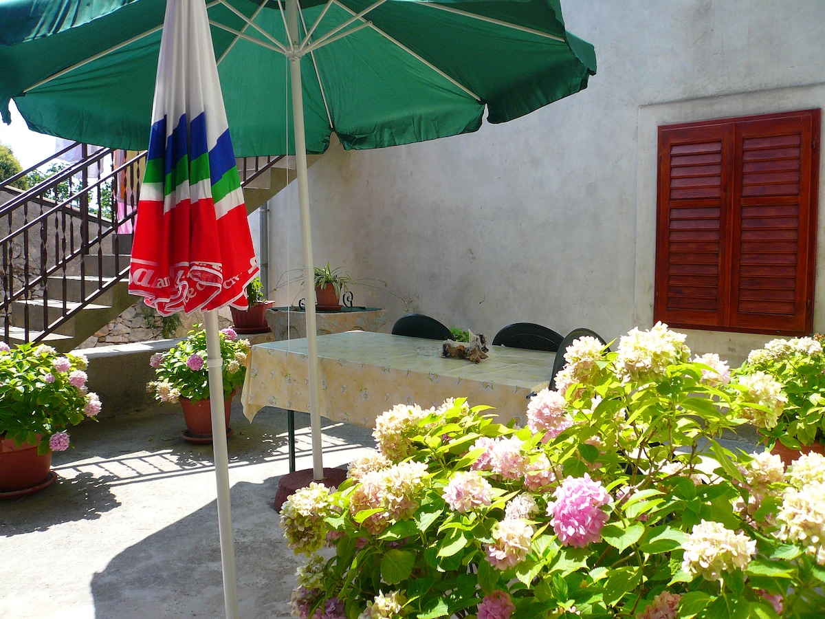 A welcoming terrace is shown, furnished with a large table and several chairs, shaded by a green umbrella. Colorful flowering plants in pots are placed nearby, adding a touch of nature. A staircase is visible in the background, leading to an upper level.