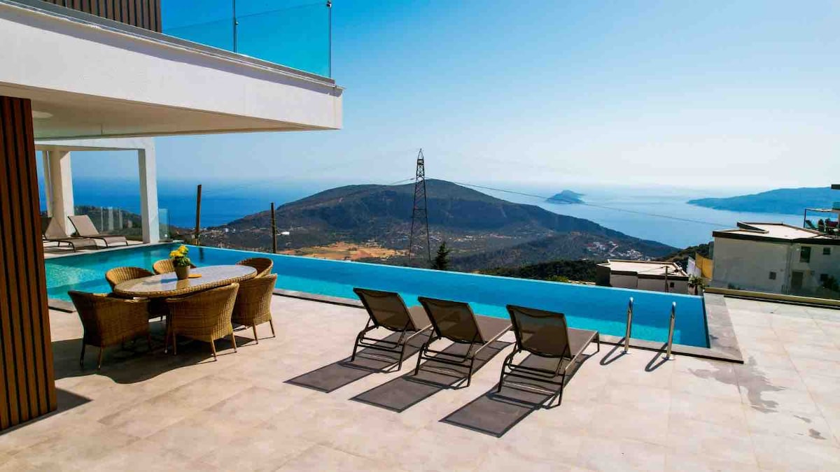 An outdoor terrace features a long, rectangular pool reflecting the sky, surrounded by four lounge chairs. A table with a vase of fresh flowers adds a welcoming touch. In the background, hills and the sea create a scenic view.