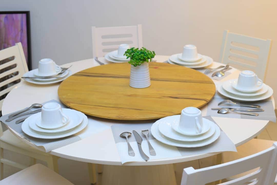 A circular dining table is set with six white plates, cups, and utensils. A wooden centerpiece adds warmth. Each place setting features a neatly arranged fork, knife, and spoon beside a cloth napkin, creating an inviting space for meals.