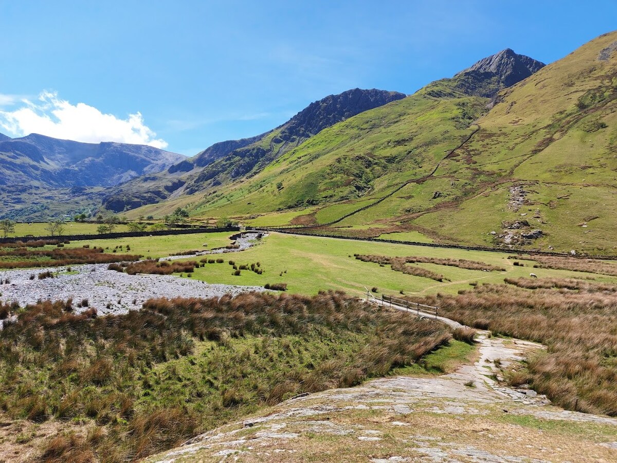 A broad expanse of green fields is framed by rising hills and mountains under a clear blue sky. Gentle slopes lead towards more rugged terrain in the distance, with patches of grass and sparse vegetation dotting the landscape.