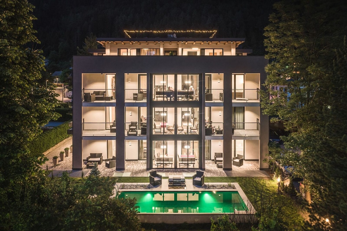 A modern building features multiple balconies and large glass doors, showcasing outdoor spaces. A serene natural pool is visible in the front, surrounded by lounge seating. The building is illuminated warmly against the backdrop of lush greenery and distant hills.