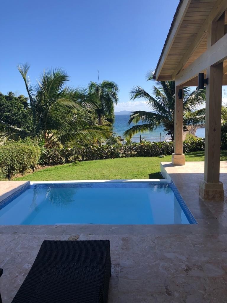 A private outdoor area features a small pool adjacent to a well-maintained lawn. Tropical palm trees and lush greenery frame the space, while a view of the marina and distant mountains is visible under a clear blue sky.