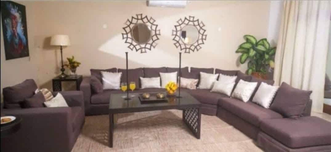 A spacious living area features a large sectional sofa adorned with various cushions. A glass coffee table sits in the center, accompanied by two tall candlesticks. Light flows in through sheer curtains, while decorative mirrors enhance the room's modern design.