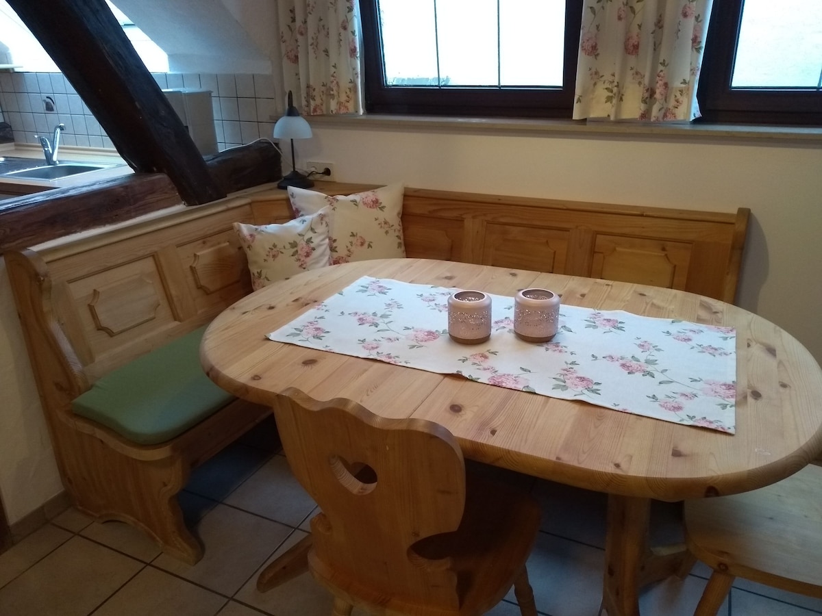 A wooden dining area features a round table set with floral linens and two decorative candles. Surrounding benches provide comfortable seating, while cheerful cushions enhance the space. Large windows adorned with floral curtains allow natural light to fill the room.