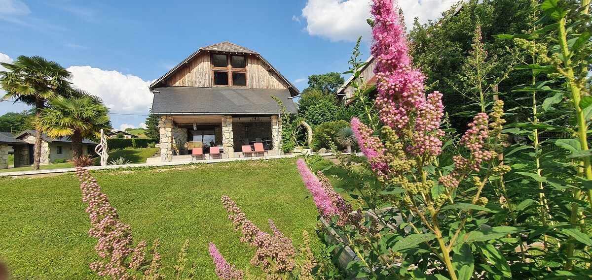 A rustic farm structure features a sloped roof and large windows, surrounded by greenery. A well-tended garden includes tall pink flowers and shrubs in the foreground, while seating areas are visible on the terrace, inviting relaxation.