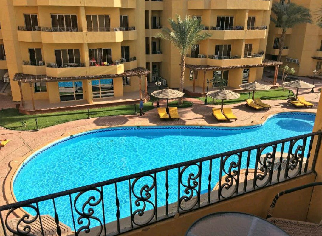 A view of a swimming pool surrounded by lounge chairs and palm trees. The pool's bright blue water is complemented by nearby shaded seating areas with yellow canopies. Surrounding balconies provide a glimpse into the cheerful architecture of the buildings.