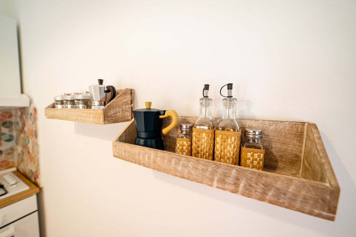 Wooden shelves are mounted on a wall, displaying a variety of kitchen items. A traditional espresso maker is positioned beside glass bottles filled with liquids, alongside several small containers. The setup offers a functional and organized kitchen space.