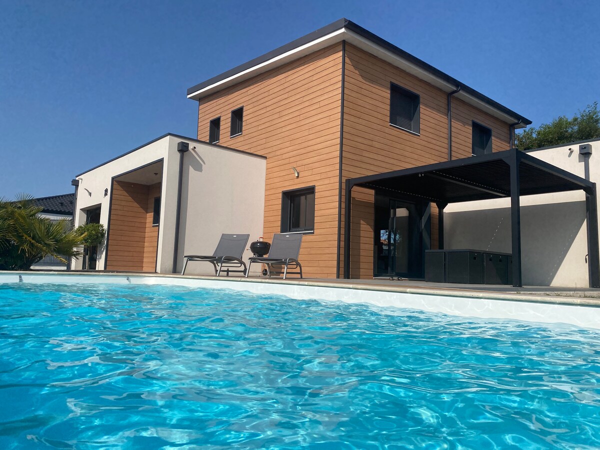 A two-story house is depicted with a wooden facade and light-colored walls. A patio area with a pergola is visible, accompanied by two lounge chairs. The foreground features a clear swimming pool, reflecting the blue sky.