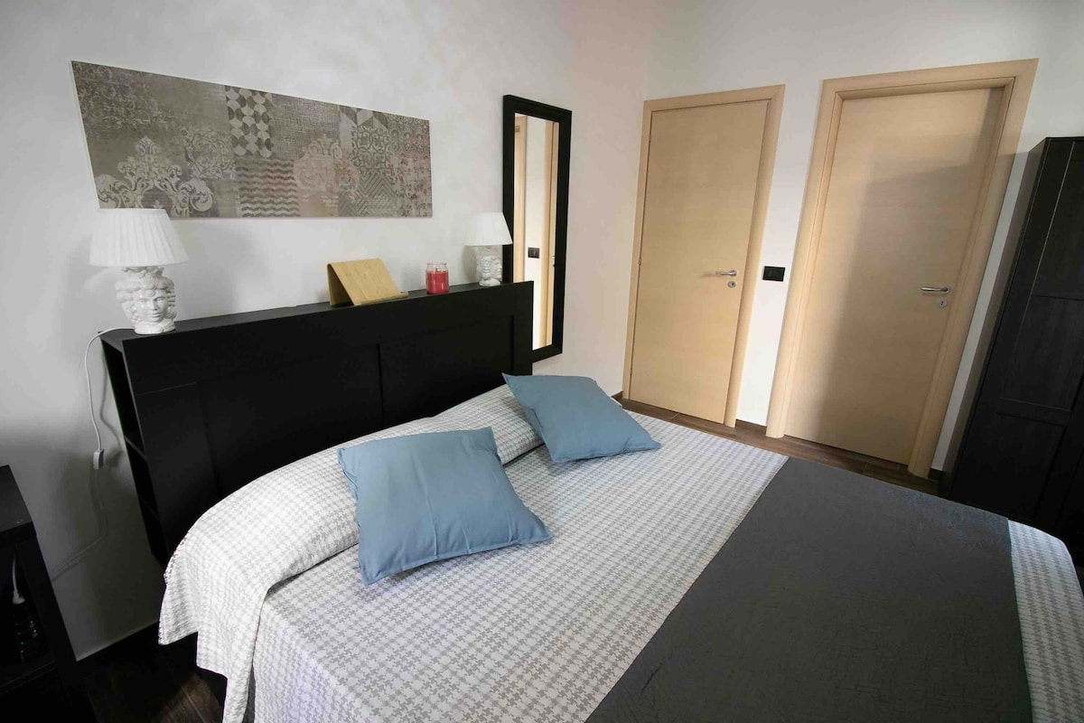 A spacious sleeping area is featured, showcasing a large bed with a light-colored bedspread and soft blue decorative pillows. Two doors are visible in the background, along with a mirror and a small decorative lamp on either side of the bed.