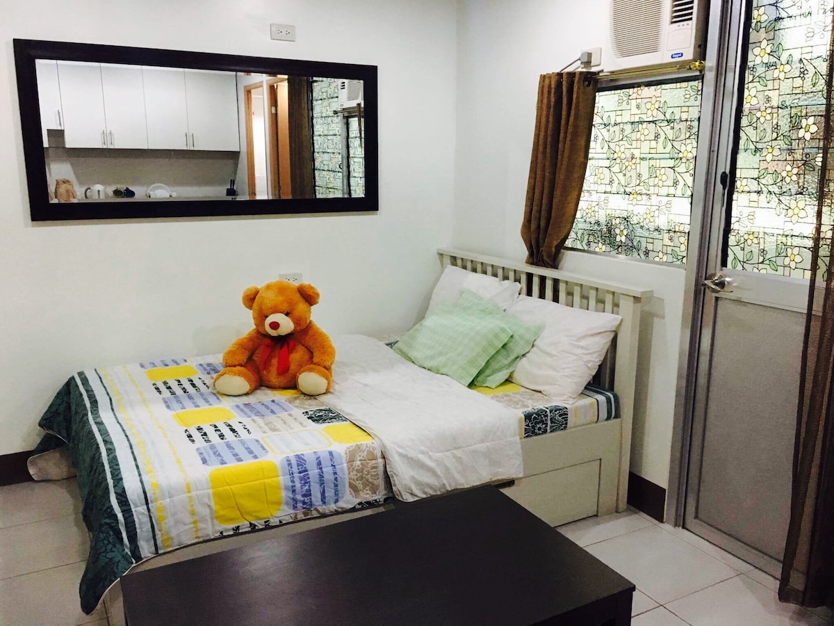A cozy sleeping area features a bed adorned with a striped blanket and a plush teddy bear. A mirror reflects the light from the nearby window with patterned curtains. The room includes a small table and cabinetry visible in the background.