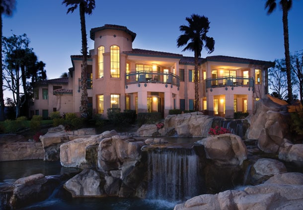 A spacious resort building is set against a twilight sky, framed by palm trees. Natural stone formations and a waterfall create a serene ambiance in the foreground. Soft lighting emanates from the large windows, highlighting the architectural features of the structure.