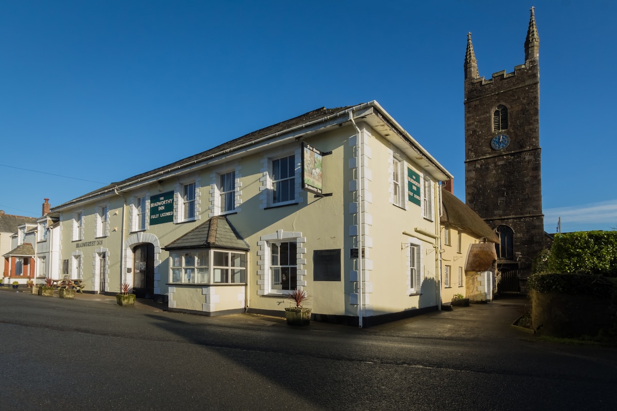 A traditional 16th century inn is depicted, featuring a yellow and white exterior. The building includes multiple windows, a small sign above the entrance, and planters with greenery. A tall stone tower with a clock is visible nearby, contributing to the historic charm of the area.