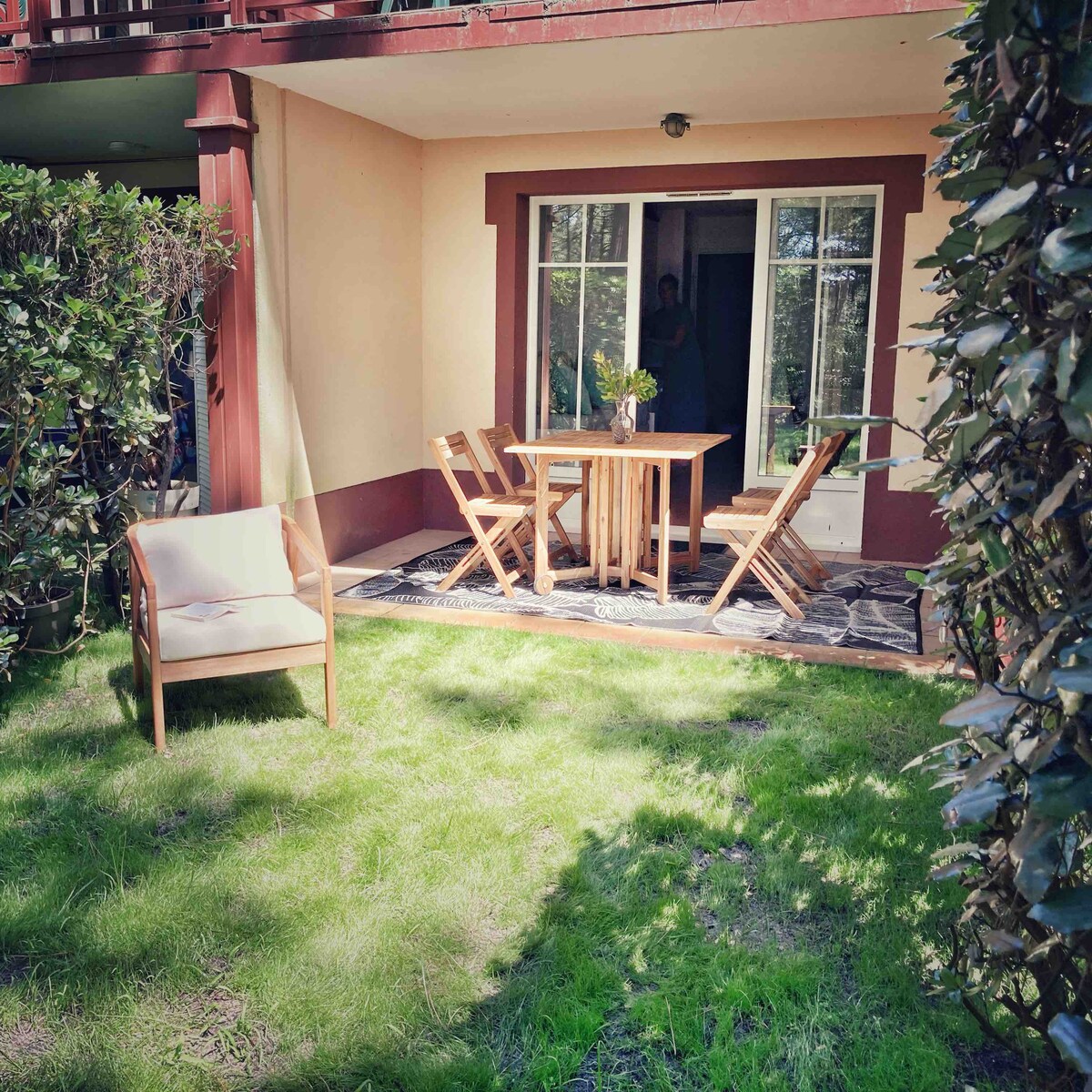 An outdoor terrace features a small table and two folding chairs, set on a grassy area surrounded by low hedges. A door provides access to the interior of the apartment, with a gentle light filtering through the nearby foliage.