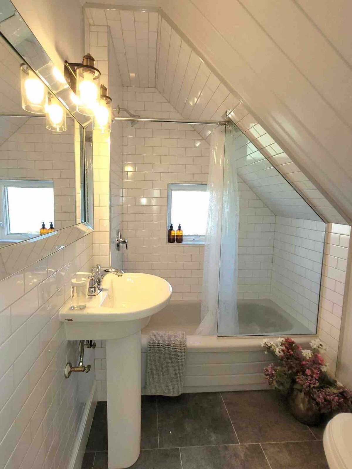 A modern bathroom features white subway tiles and an angled ceiling. A glass shower enclosure is positioned beside a soaking tub. Natural light enters through two small windows. A pedestal sink is present, accompanied by stylish lighting fixtures and decorative plants by the tub.