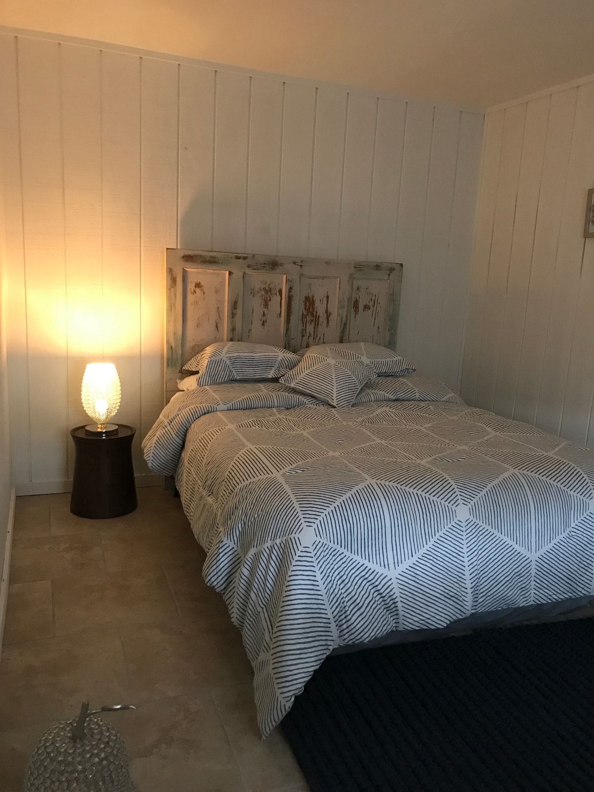 A cozy sleeping area features a bed with a patterned blanket and two pillows. A small modern lamp provides soft light on a side table. The room is decorated with white walls and neutral tile flooring, creating a calm and simple atmosphere.