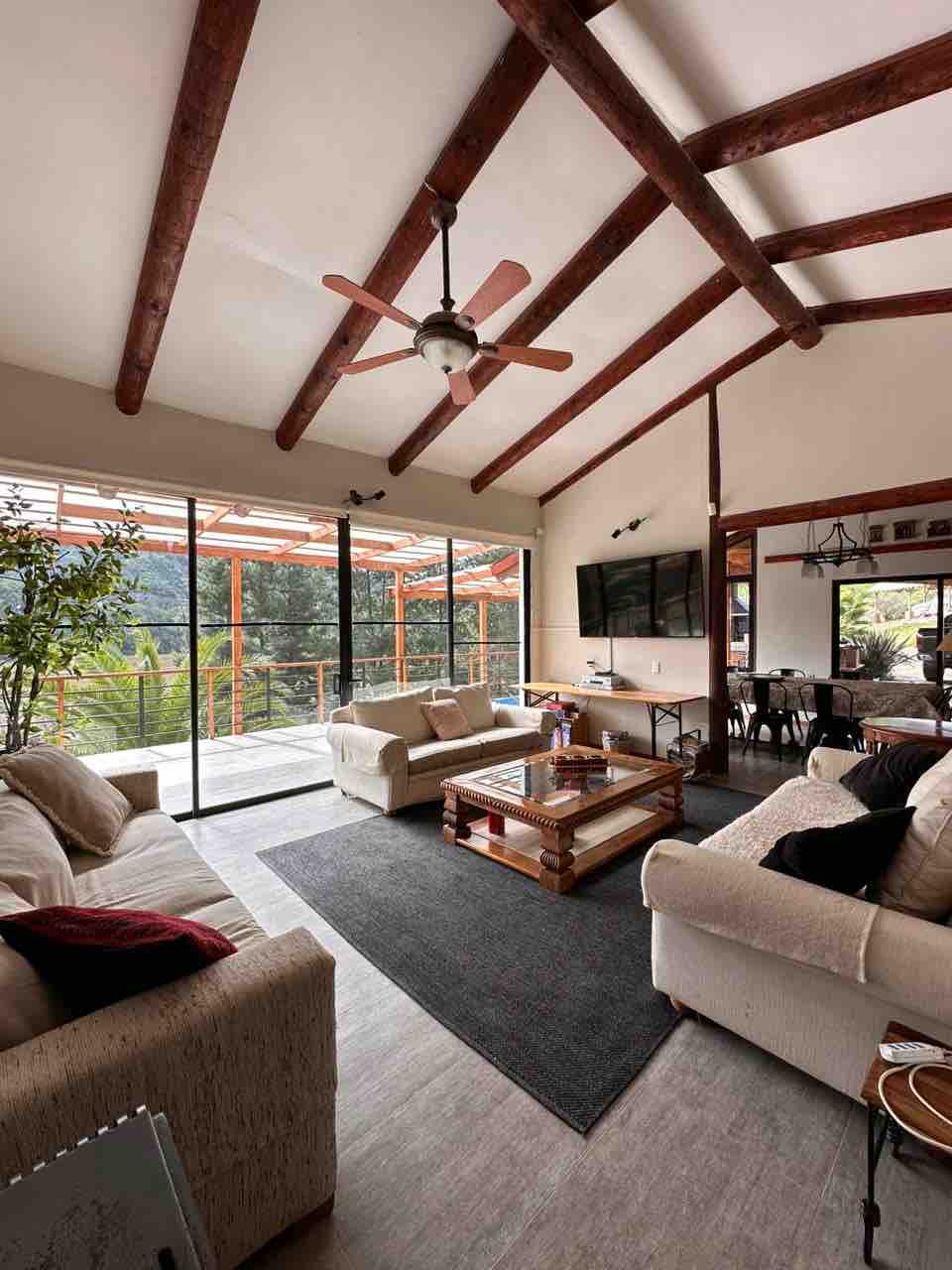 A spacious living area features high wooden beams and large glass doors that open to a scenic outdoor view. Comfortable sofas are arranged around a wooden coffee table, and a flat-screen television is mounted on the wall. A dining space is visible in the background.