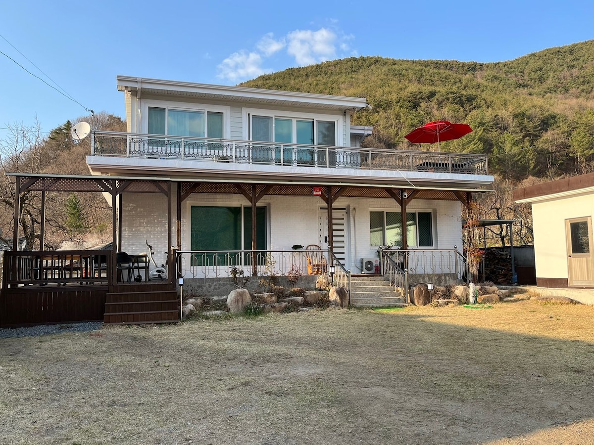 The two-story house features a large balcony with an umbrella, surrounded by a grassy area. Glass doors provide ample natural light to the interior. A staircase leads to the entrance, while natural greenery is visible in the background, contributing to a serene setting.