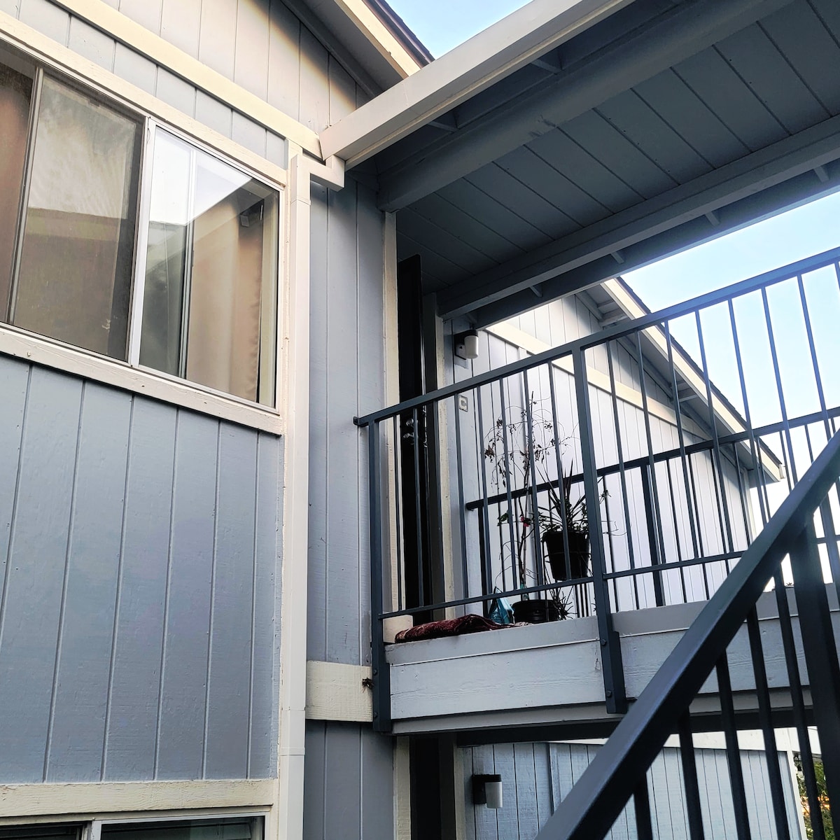 The exterior features a two-story structure with a blue and gray color palette. A staircase leads up to a small balcony, which includes a railing and a potted plant. Windows on the upper floor allow natural light to enter.