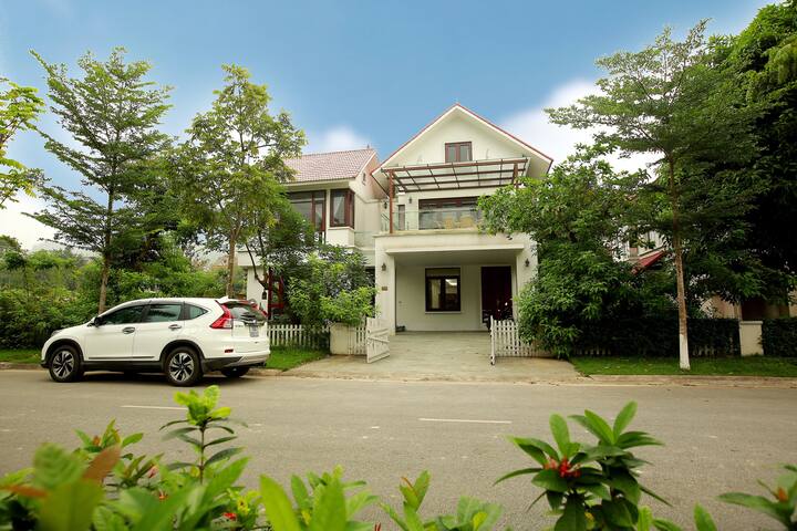 A modern villa is displayed, featuring a spacious façade with large windows and a covered entryway. The surrounding greenery includes trees and shrubs, creating a serene atmosphere. A white car is parked in the driveway, enhancing the villa's welcoming appearance.