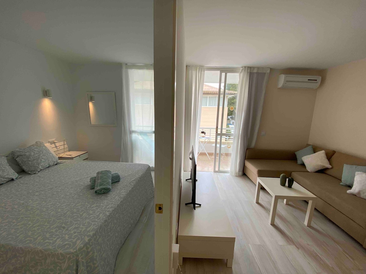 A bright and airy living space is divided into sleeping and relaxation areas. A spacious bed is complemented by soft linens and towels. The lounge features a comfortable sofa and a coffee table, with large glass doors that open to an outdoor seating area, allowing natural light to fill the room.