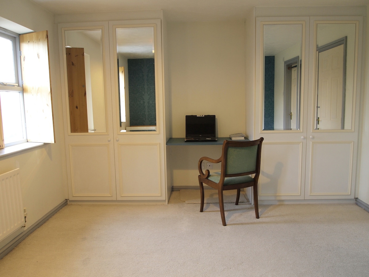 A spacious room is presented with light-colored walls and a soft carpet. A desk with a laptop is positioned against the wall, accompanied by a vintage chair. Two sets of sliding mirror doors are visible, adding depth to the space, while a window allows natural light to enter.