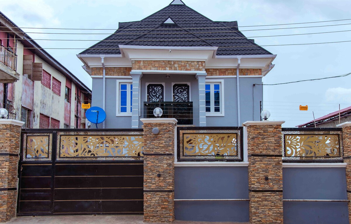 The exterior of the apartment is characterized by a modern design, featuring a combination of light grey walls and decorative stone accents. The entrance is framed by ornate metalwork gates, and large blue windows allow natural light in. A satellite dish is visible on the side.