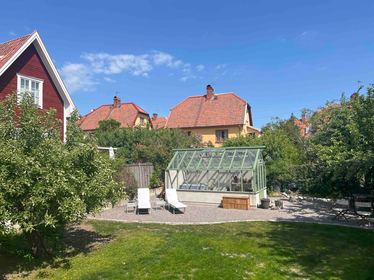 A garden area is showcased with a greenhouse surrounded by foliage and spacious gravel pathways. Two loungers are positioned on the lawn, with several trees providing shade. Nearby, colorful houses with red roofs are visible under a clear blue sky.