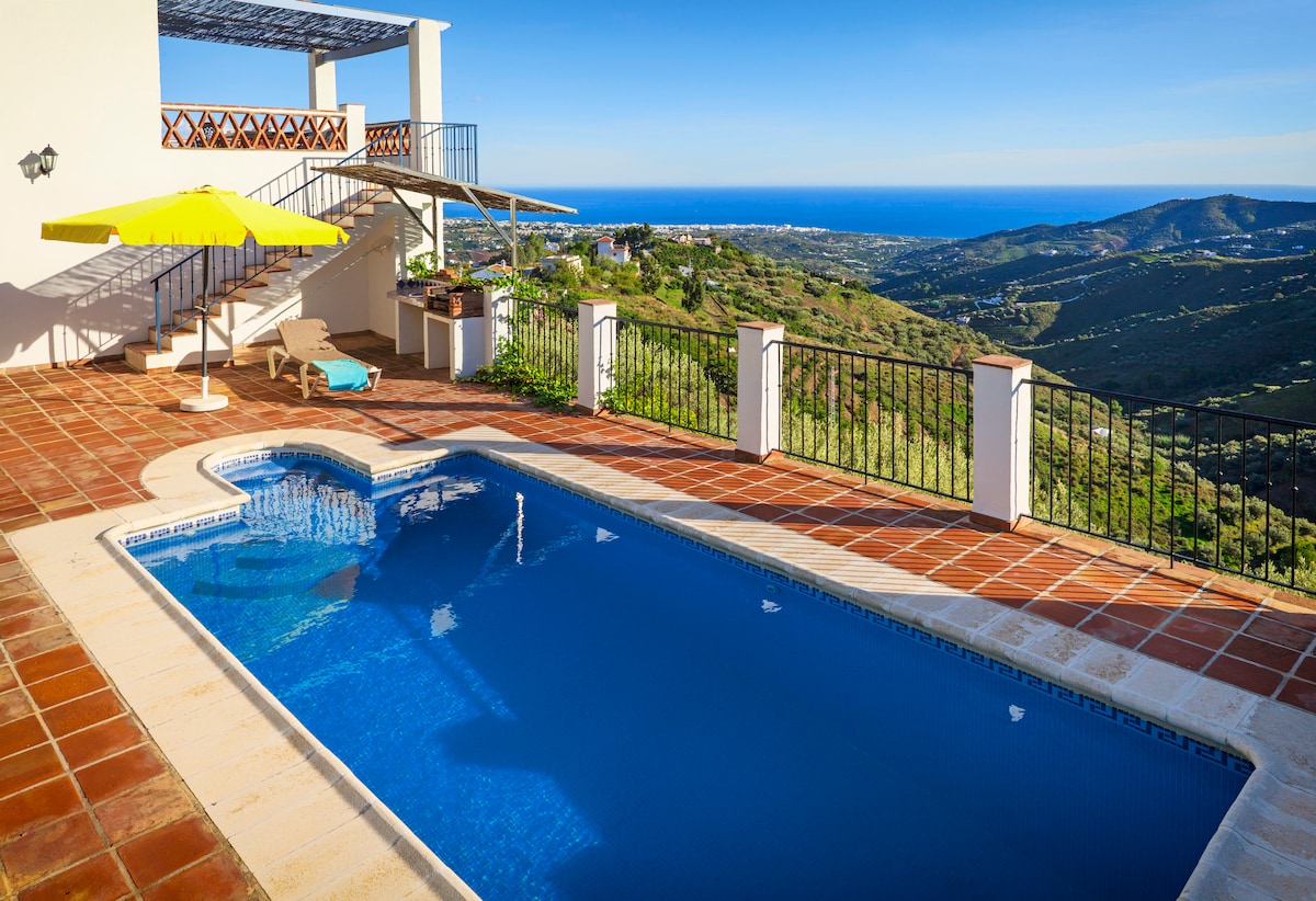 A private swimming pool is set against a backdrop of rolling hills and distant sea views. A sun umbrella and lounge chair are positioned nearby, inviting relaxation on the sun-drenched terrace. The area features stylish railing and terracotta tiles that enhance the outdoor space.