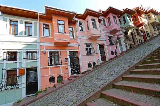 A row of charming, colorful buildings is viewed from the bottom of a steep cobblestone street. The architecture features rectangular windows, decorative grilles, and distinct pastel colors, creating a lively streetscape that reflects the historical character of the Fener district.