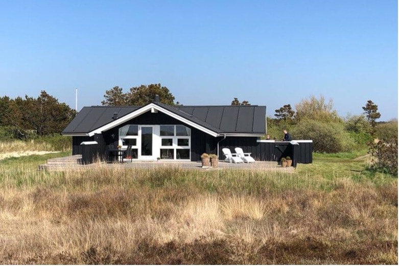 A modern holiday home is set against a clear blue sky, featuring a dark facade and large windows. The wide wooden deck is equipped with white lounge chairs and offers views of the surrounding natural landscape. Tall grasses and sparse vegetation are visible in the foreground.