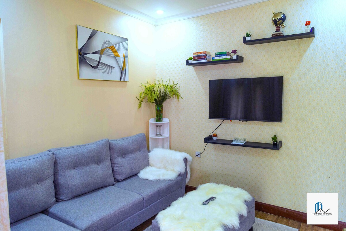 A comfortable seating area features a gray sectional sofa with a fluffy white throw. Above, a mounted flat-screen TV is complemented by simple black shelves displaying a few books and decorative plants. Warm light highlights the soft textures and welcoming ambiance.
