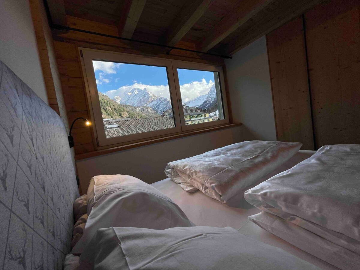 A serene sleeping area features a spacious bed adorned with fluffy pillows. Large windows frame a stunning mountain view, showcasing peaks that are partially covered in snow. Soft natural light illuminates the room, highlighting wooden accents in a peaceful setting.