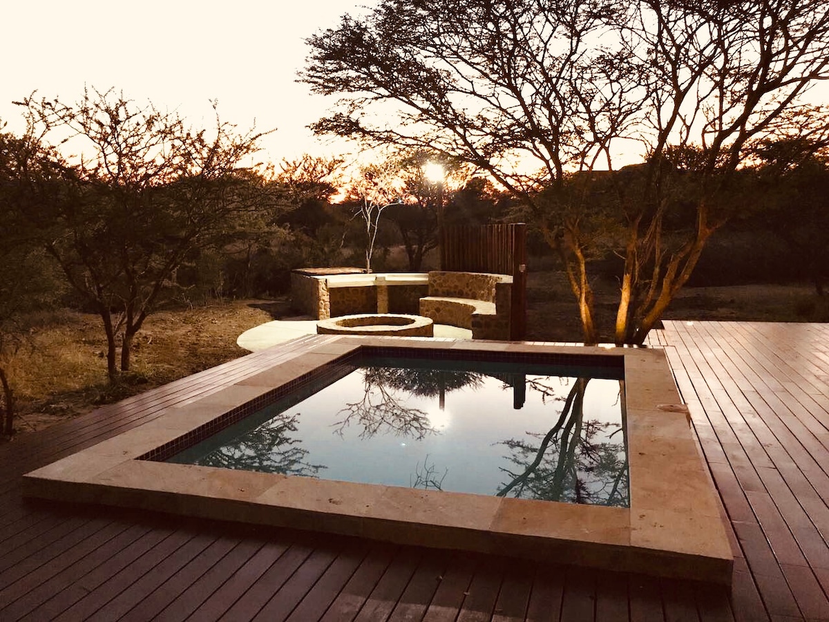 A private swimming pool is set on a wooden deck, surrounded by native trees. The water reflects the evening sky, while a boma area with seating is nearby. Soft lighting adds warmth, creating a serene outdoor space suitable for relaxation and gatherings.