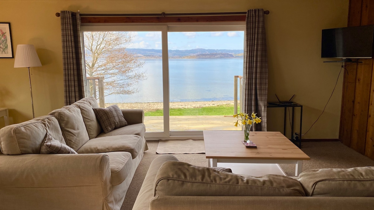 A comfortable seating area is presented with two light-colored sofas, facing large sliding glass doors that open to a view of Loch Fyne. A wooden coffee table sits at the center, adorned with a small vase of yellow flowers. Soft natural light fills the room.