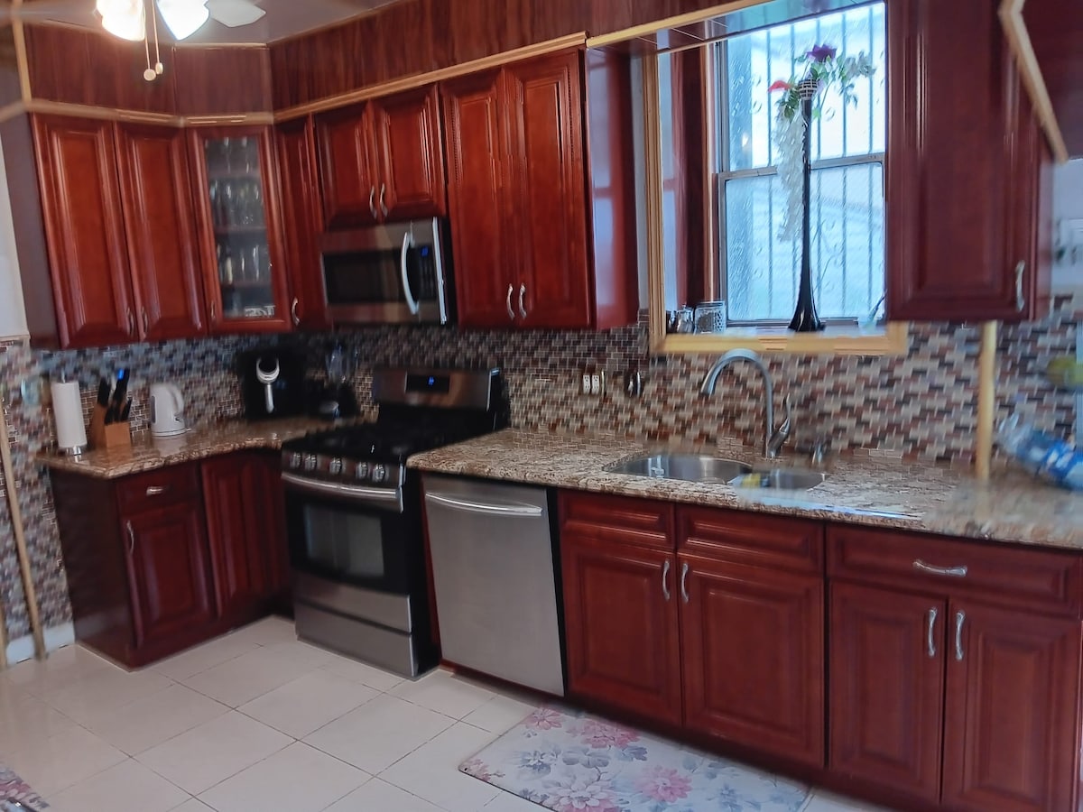 A spacious kitchen features wooden cabinets and granite countertops. Modern stainless steel appliances, including a fridge, stove, and dishwasher, are positioned for convenience. A decorative vase with flowers sits on the windowsill, while patterned tiles add texture to the backsplash and floor.