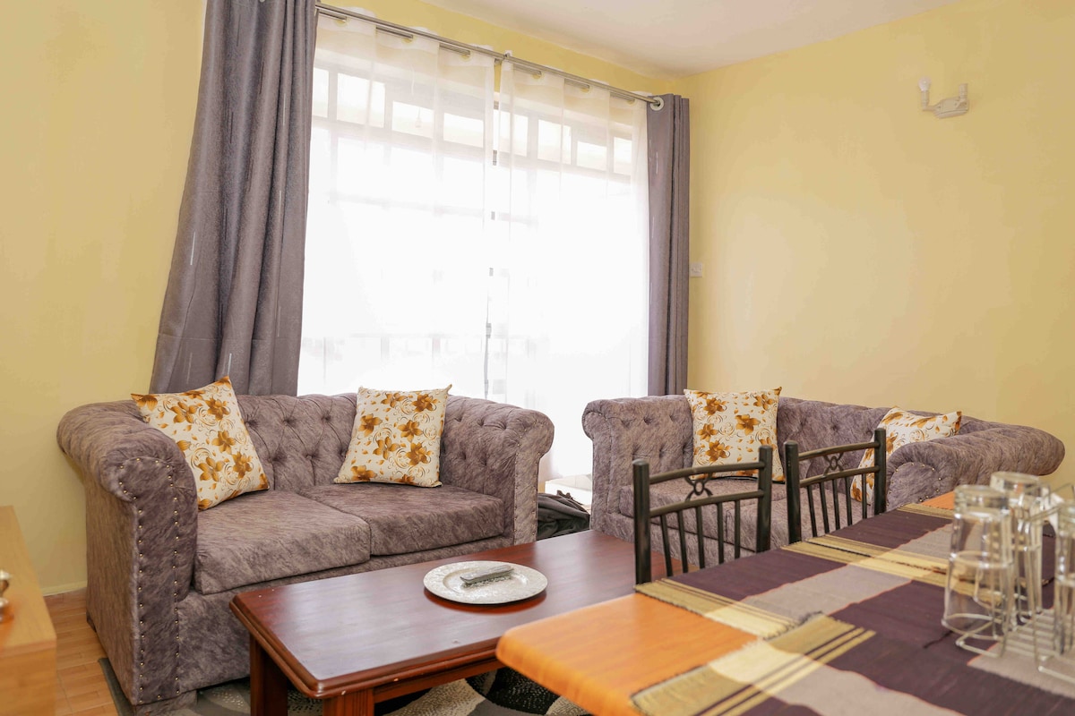 A living area is presented with two plush sofas adorned with floral-patterned cushions. A wooden coffee table is positioned in front, and a dining table featuring dark chairs is visible to the side. Natural light filters through sheer curtains, brightening the space.