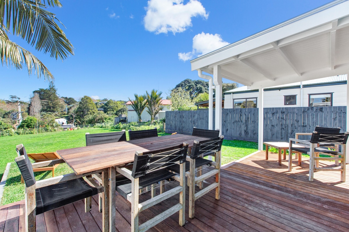 An outdoor deck area features a large wooden dining table surrounded by black chairs. The space is complemented by well-kept grass and a fence in the background. Blue skies and a few clouds enhance the serene outdoor setting.