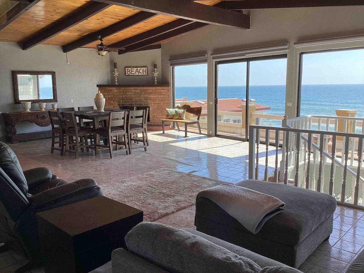 An inviting living space features large windows offering views of the ocean. A dining table with seating for several is positioned near the light-filled area. Comfortable seating arrangements include a sofa and armchairs. Natural light complements the earthy tones and wooden beams of the ceiling.